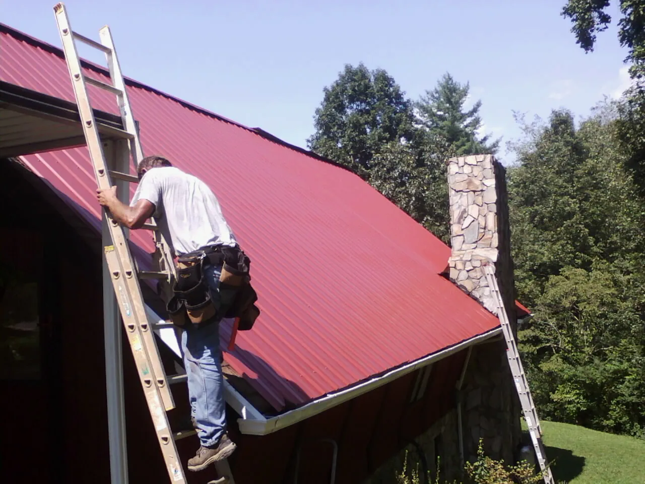 Roofer on a ladder beside a red metal roof and stone chimney — Soffit & Fascia Repair by Roofing Brothers in Bergenfield